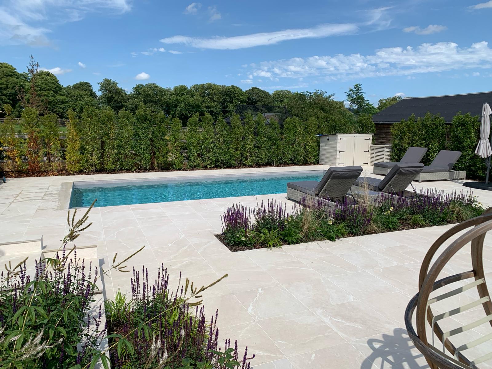 Outdoor swimming pool area with light stone paving, sun loungers, and purple flower beds.